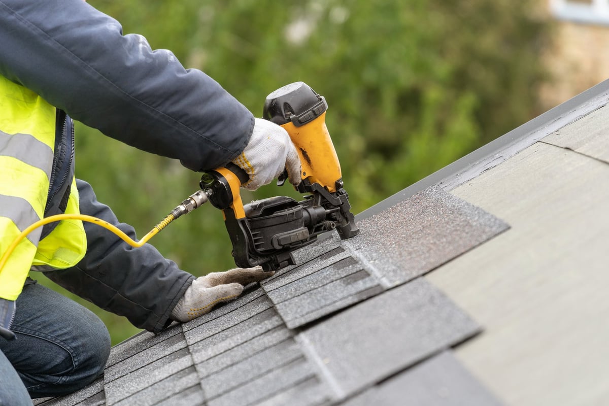 Unrecognizable roofer worker using pneumatic nail gun installing asphalt shingles on a residential roof under construction
