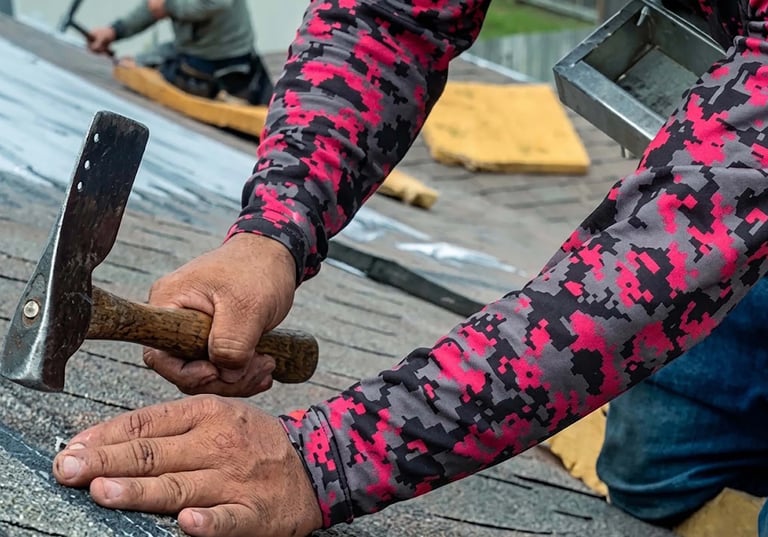 Person wearing pink and black patterned sleeves hammering nails into roofing material on a construction site